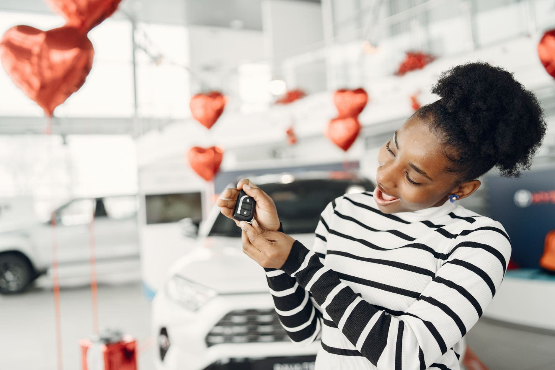 Joyful woman holding car key amidst heart decorations in a showroom.